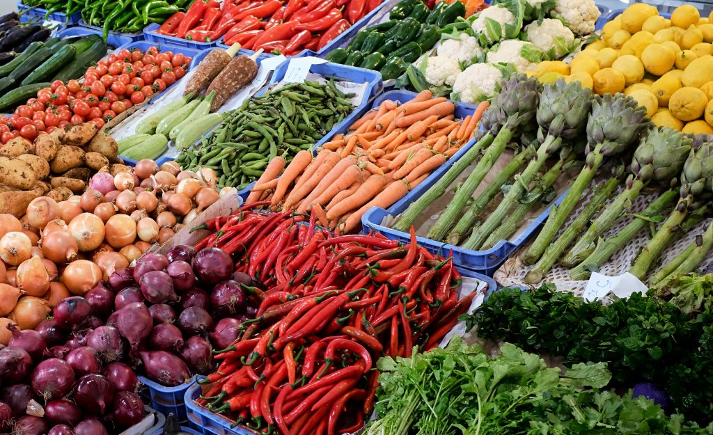 zdjęcie przedstawia różne warzywa używane w diecie śródziemnomorskiej, rozstawione na targowisku, w skrzynkach leżą między innymi kolorowe cebule, karczochy, kalafior, kolorowe papryki, cytryny