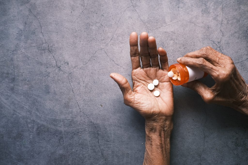 zdjęcie przedstawia pomarszczone dłonie; na lewej leżą białe tabletki, prawa trzyma pojemnik, z której wysypuje lekarstwa