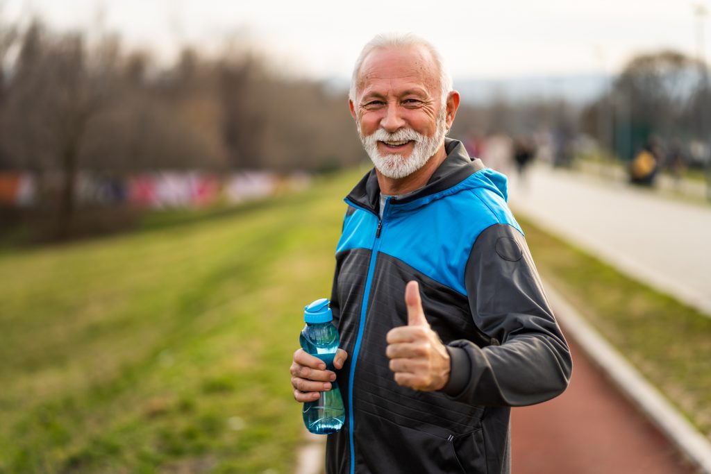 zdjęcie przedstawia mężczyznę w wieku emerytalnym, który trzyma w dłoni bidon z wodą; jest w trakcie aktywności fizycznej, prawdopodobnie joggingu