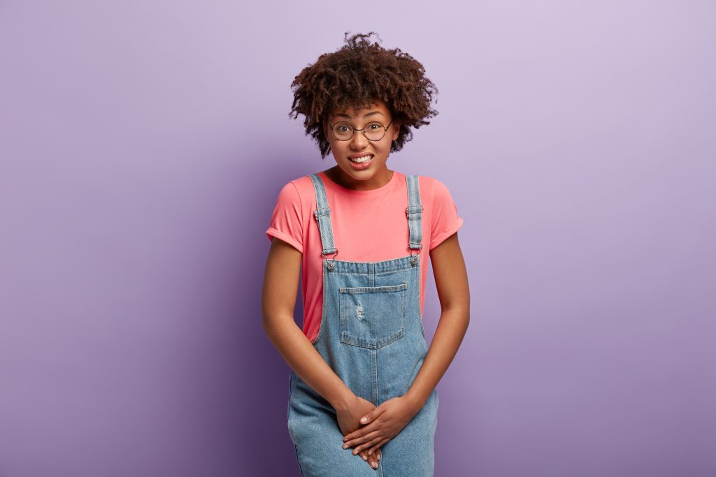 Unhappy dark skinned woman holds crotch, needs toilet, has problematic situation, wears pink t shirt and denim sarafan, suffers from cystitis, isolated over purple background. People and urgency