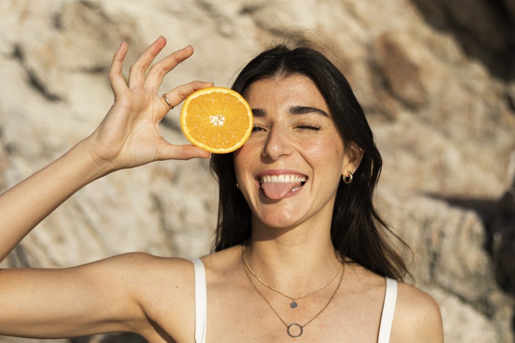 zdjęcie: uśmiechnięta młoda dziewczyna trzyma połówkę pomarańczy na wysokości prawego oka; ma zawadiacką minę i wystawiony język. Osoba stoi na słońcu i prawdopodobnie uzupełnia braki w witaminie d
