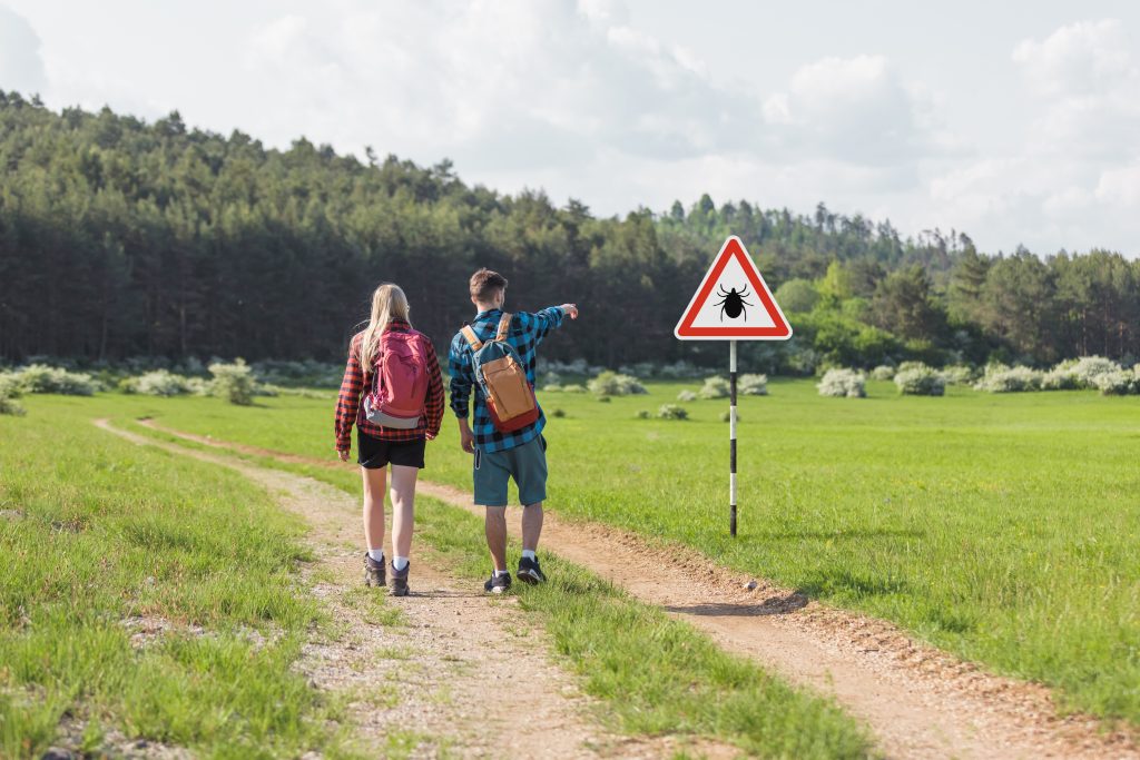 zdjęcie przedstawia parę: chłopaka i dziewczynę w czasie trekkingu, chłopak ręką wskazuje na znak ostrzegający przed kleszczami