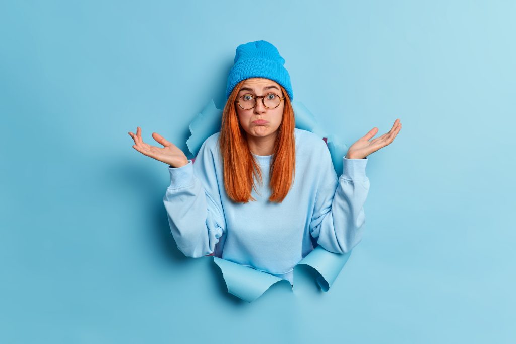 Confused puzzled red haired young questioned female shrugs shoulders and spreads palms sideways stands clueless in ripped hole of blue paper background wears trendy hat casual jumper cannot decide.