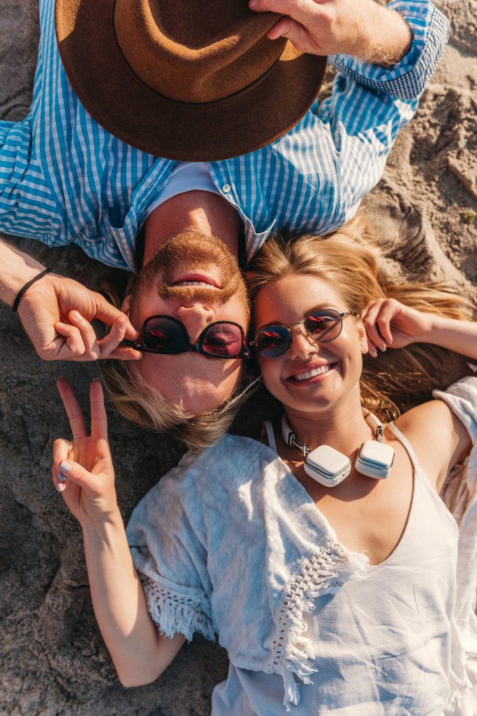 view from above on young attractive smiling happy man and woman in sunglasses lying on sand beach, romantic couple by the sea on sunset, boho hipster style outfit, friends having fun together