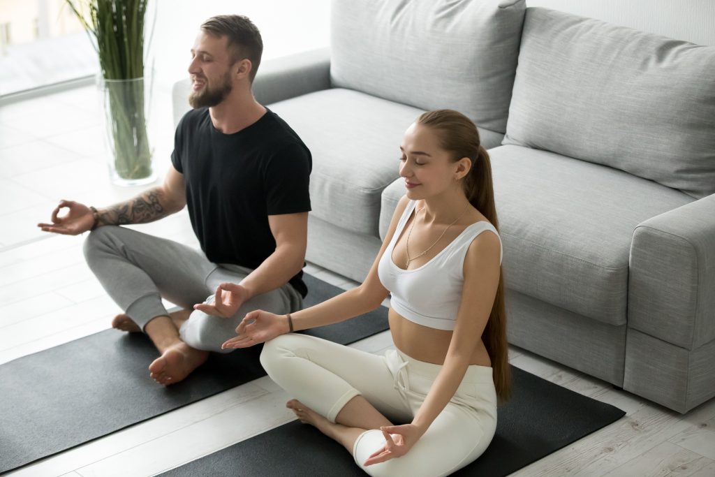 Calm smiling man and woman sit on fitness mats on floor meditating together, happy millennial couple train together practicing yoga at home, boyfriend and girlfriend doing exercises in lotus position