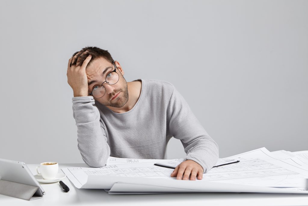 Tired male illustrator being fatigue after drawing for long time. Attractive bearded man in spectacles scratches head in perplexity, has some troubles with future project. Overworked architect