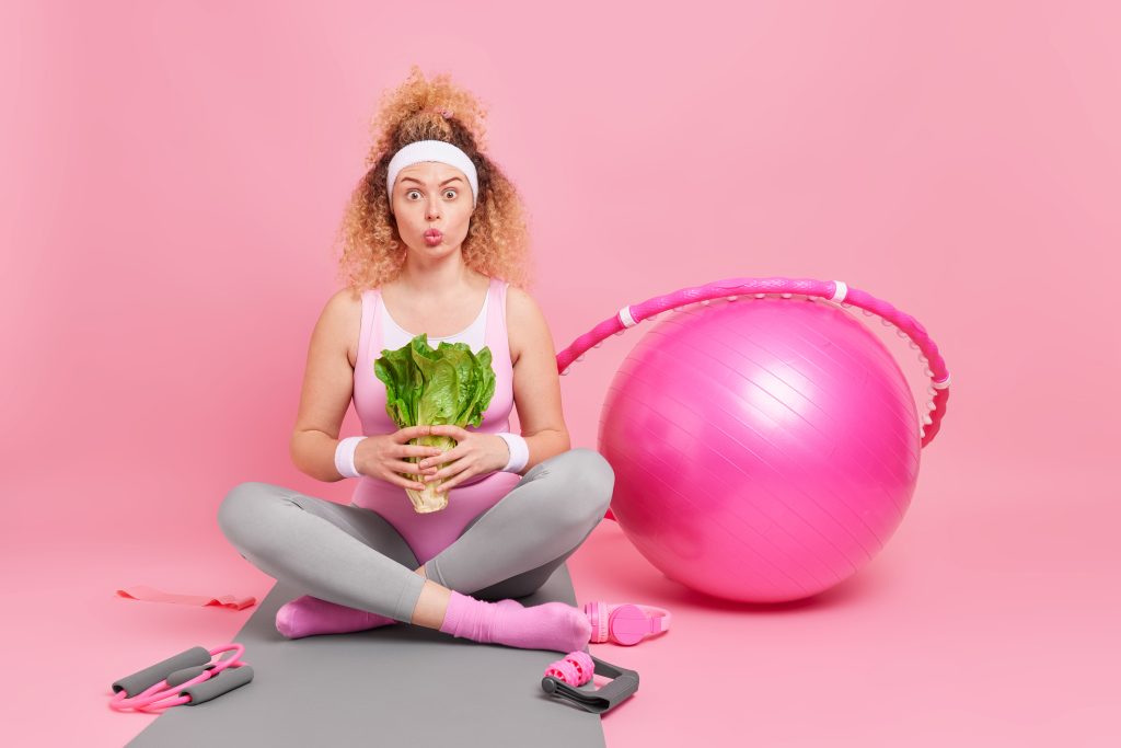 Photo of surprised curly woman holds green vegetable has healthy nutrition and active lifestyle sits crossed legs on fitness mat exercises at home isolated on pink wall. Sport dieting wellness concept