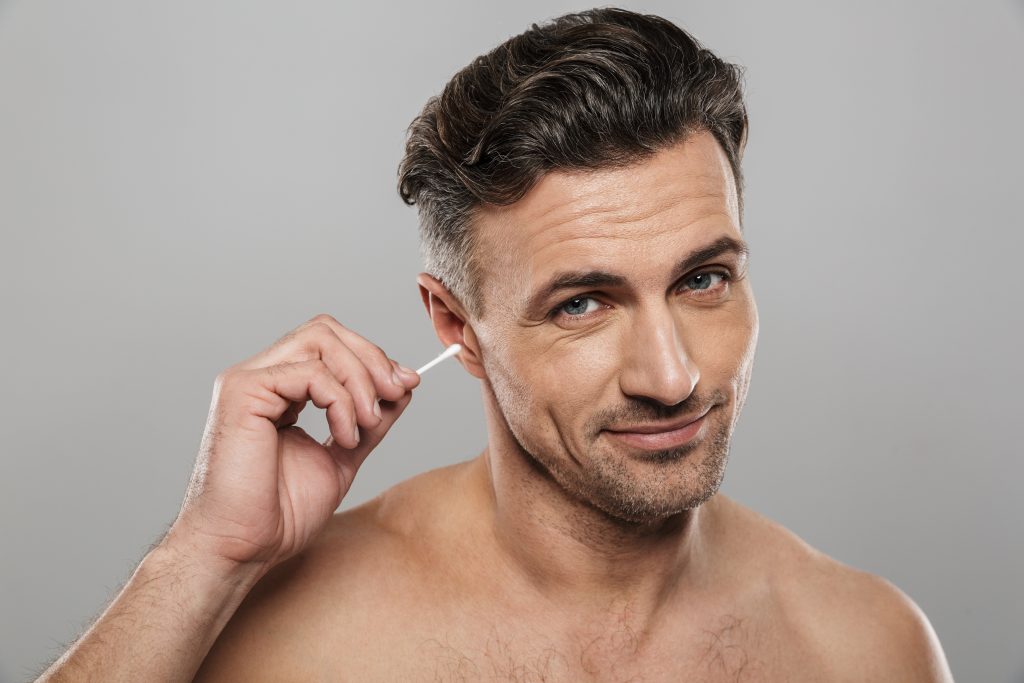 Image of handsome mature man standing isolated over grey wall background naked holding cotton bud take care of his ears cleaning it.