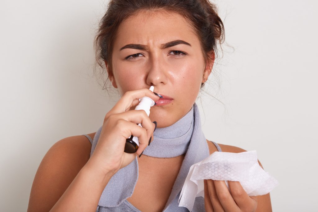 Horizontal indoor studio shot of bad looking woman having caught cold, feeling unwell, being on sick leave, using nose spray, holding napkin in one hand, running nose. People and illness concept.