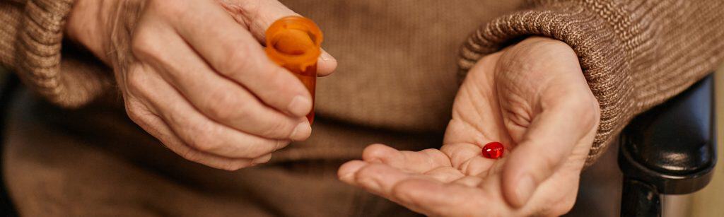 Website header shot of senior Caucasian man sitting in wheelchair holding orange pill bottle and dispensing red medication tablet into palm, close up on hands, nursing home healthcare concept
